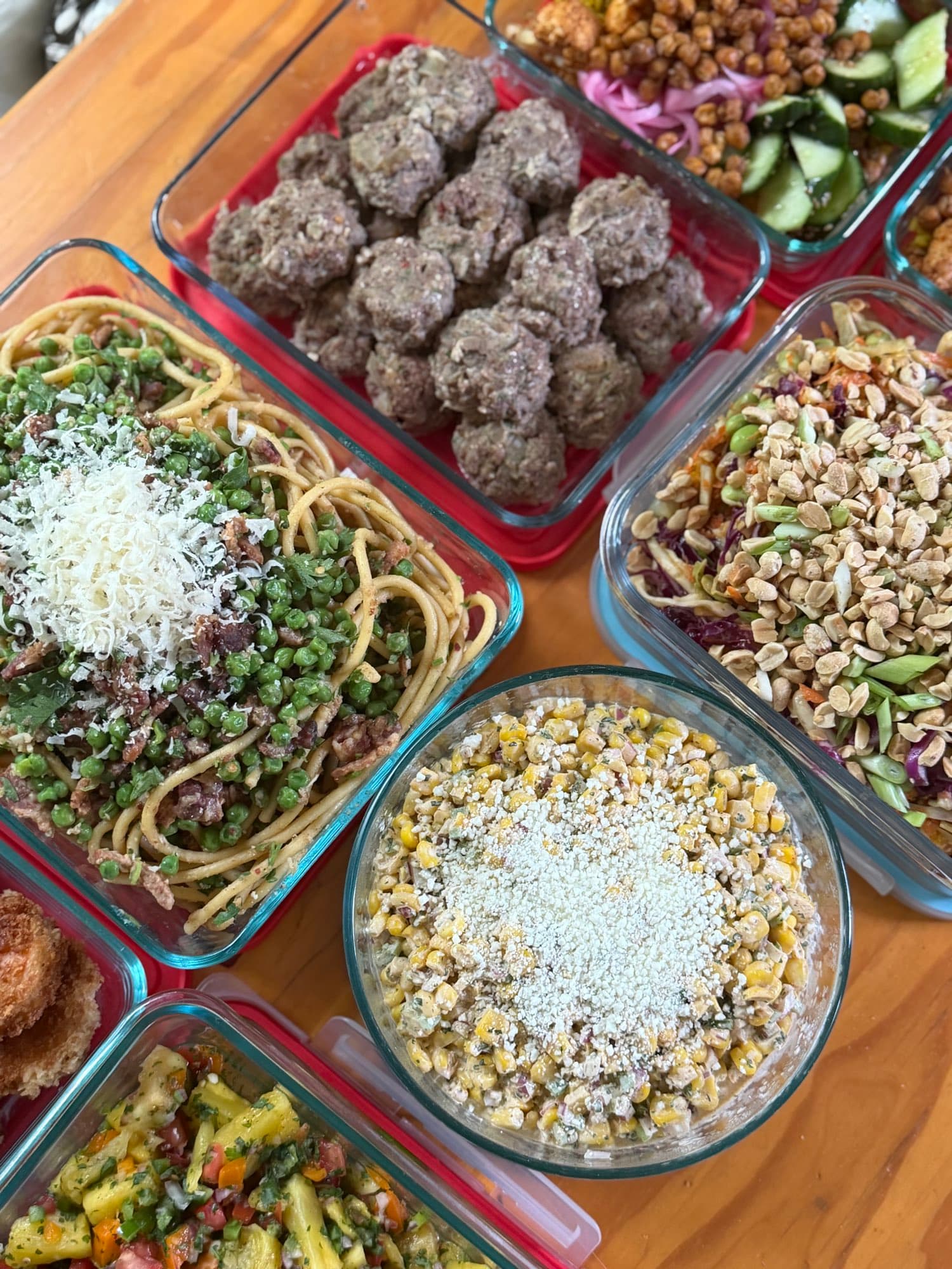 A table of prepped containers: meatballs, spaghetti with peas, crunchy slaw, elote corn salad, and mango salsa